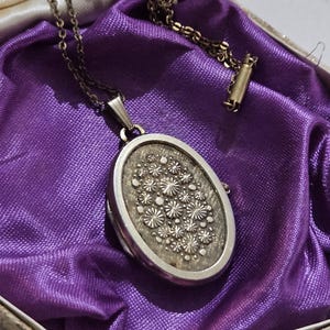 Può includere: Una collana con un medaglione ovale in argento con un motivo floreale, appoggiata su un tessuto di raso viola all'interno di una scatola color crema. La catena della collana è visibile e il medaglione è chiuso.