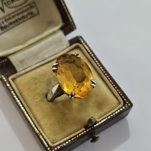 May include: A silver ring featuring a large, oval-cut, golden-yellow citrine gemstone held by four prongs. The ring is displayed inside a dark brown, hinged jewellery box with a beige velvet interior and gold-coloured trim along the edges.