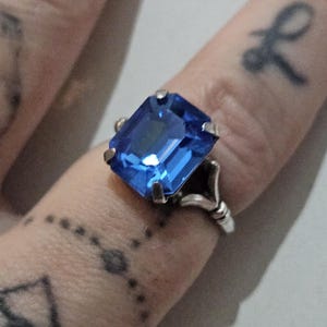 May include: A silver ring featuring a large, rectangular, brilliant blue gemstone. The ring's setting has an ornate design with delicate details. The gemstone is the focal point, catching the light and reflecting a vibrant color.