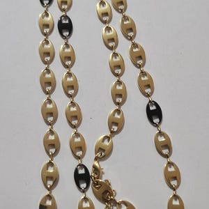 Può includere: Collana dorata con una catena di maglie ovali, intervallate da maglie ovali nere. La collana ha una chiusura a moschettone e una piccola etichetta con la scritta "GUESS". Il gioiello è esposto su una superficie bianca.
