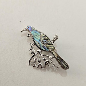 May include: A silver-toned bird brooch with iridescent blue and green accents. The bird is perched on a detailed branch. The brooch is a decorative accessory, suitable for adding a touch of nature-inspired elegance to clothing or accessories.