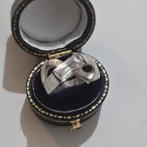 May include: A silver ring with a bow-shaped design, displayed in a black jewellery box with a blue interior. The box is round and has gold detailing.