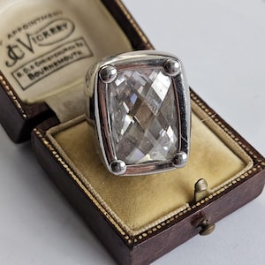 May include: A silver ring featuring a large, rectangular, faceted clear gemstone set in a four-prong mounting. The ring is displayed inside a dark brown, hinged jewellery box with a cream-coloured velvet interior and the text J.C. Vickery Bournemouth.