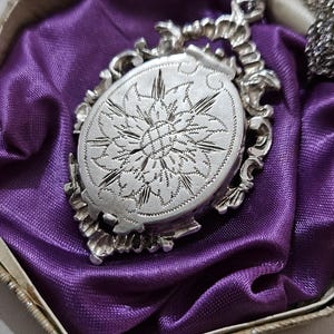 May include: A silver-toned oval pendant necklace with an ornate floral design. The pendant, with a detailed etched pattern, rests on a bed of purple satin within a vintage box. The necklace chain is also silver-toned.