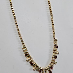 Puede incluir: Un collar de oro con una cadena delicada y un diseño en cascada de diamantes de imitación transparentes y rojos.