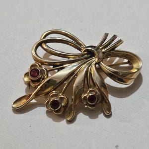 May include: A gold-coloured brooch with a bow design and three small flower clusters. Each flower has a red gemstone centre. The brooch has a vintage aesthetic and is likely made of metal.