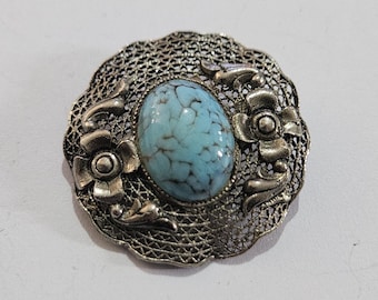 May include: A silver filigree brooch with a turquoise cabochon stone in the center. The brooch is decorated with two silver flowers on either side of the stone.