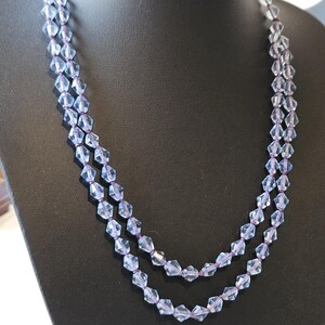 May include: A double-strand necklace featuring light blue, faceted beads. The beads are strung on a purple cord and presented on a black jewellery bust. The necklace has a delicate, elegant appearance.