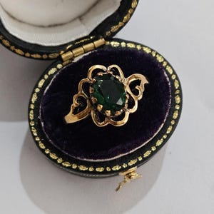 May include: A gold ring with an oval, emerald-green gemstone set in a decorative filigree design. The ring is displayed inside a black jewelry box with a purple velvet interior and gold trim.