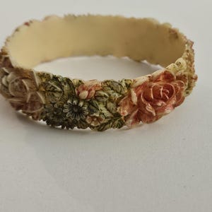 May include: Ivory-colored bangle bracelet with a floral design. The bracelet features carved roses, leaves, and other flowers in shades of peach, green, and cream. The bracelet is approximately 2.5 inches in diameter.