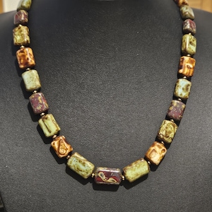 Könnte beinhalten: Eine Halskette mit rechteckigen Perlen in Grün-, Braun- und Burgundertönen, die durch goldfarbene Verbinder verbunden sind. Die Halskette wird auf einer schwarzen Schaufensterpuppe präsentiert.