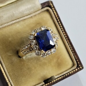 May include: A gold ring featuring a large, rectangular, deep blue centre stone surrounded by a halo of small, clear, round-cut stones. The band is also set with small, clear, round-cut stones along the sides, presented in a dark brown box.