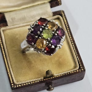 May include: A silver ring featuring a cluster of nine round, multi-colored faceted gemstones, including red, yellow, purple, and green stones, arranged in a square pattern. The ring is displayed inside a brown velvet-lined jewelry box.