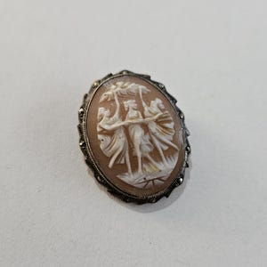 Puede incluir: Broche de camafeo ovalado con un detallado relieve de tres figuras con túnicas fluidas. El camafeo está engastado en un marco plateado con un borde decorativo. El fondo es de color melocotón claro.