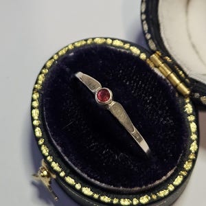 May include: A silver ring featuring a small, round, red gemstone. The ring is presented in an open, black jewellery box with a dark blue velvet interior and gold detailing.