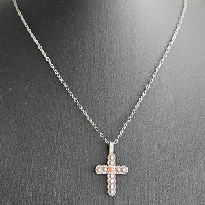 Può includere: Una collana con ciondolo a croce in metallo color argento con una piccola pietra rotonda di colore arancione incastonata al centro. La croce presenta un motivo testurizzato simile a fiori lungo i bordi ed è appesa a una sottile catena.