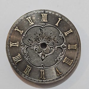以下が含まれることがあります： 金色のローマ数字と装飾的な花のセンターを備えた、アンティークシルバー調の時計の文字盤。時計の文字盤は円形で、テクスチャード加工された背景があります。金色の数字と花の装飾が特徴です。針はありません。