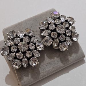 May include: A pair of silver-toned clip-on earrings. Each earring features a cluster of clear, faceted rhinestones in various shapes and sizes, arranged in a floral pattern. The earrings are displayed on a grey surface.