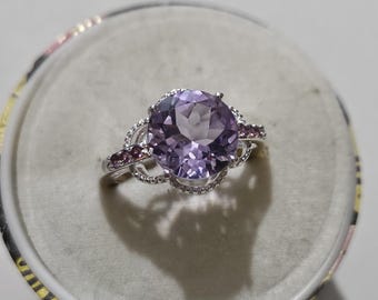 May include: A silver ring with a large, round, light purple gemstone as the centerpiece. Smaller, darker purple stones are set on the sides. The ring is displayed on a light gray surface.