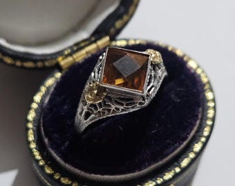 May include: A vintage ring with a square, amber-colored gemstone set in a filigree silver band. The ring is displayed in an open, black jewelry box with a purple velvet interior and gold trim. The ring's design includes small gold accents.