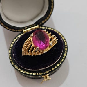 Op de afbeelding: Een gouden ring met een grote, ovale, roze edelsteen, gezet in een decoratieve gouden band. De ring wordt gepresenteerd in een open, zwart fluwelen sieradendoosje met gouden rand. De doos is open en onthult de ring.