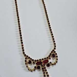Pode incluir: Um colar de ouro com um pingente de laço de cristal bordô. O colar é feito de pequenos cristais redondos e tem uma corrente delicada.