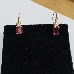 May include: A pair of gold drop earrings featuring rectangular, deep red faceted gemstones. Each earring has a small, decorative gold setting above the stone and a curved wire hook for attachment, displaying a classic and elegant design.