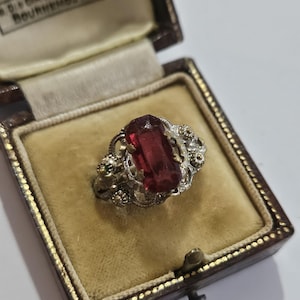 Può includere: Un anello con una pietra preziosa rettangolare sfaccettata rosso scuro, incastonata in una fascia di metallo argentato con intricati dettagli in filigrana, presentato all'interno di un portagioie marrone con rivestimento in velluto color crema.