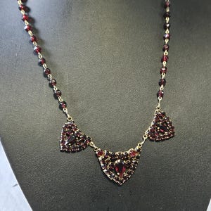 Puede incluir: Un collar vintage con colgantes en forma de corazón y una cadena de pequeñas cuentas facetadas de color rojo oscuro. El collar se exhibe en un maniquí negro. El metal parece ser de color dorado.