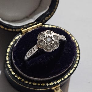 May include: A silver ring featuring a floral cluster of small, round gemstones. The ring is presented in a black jewellery box with a dark blue velvet interior and gold detailing.