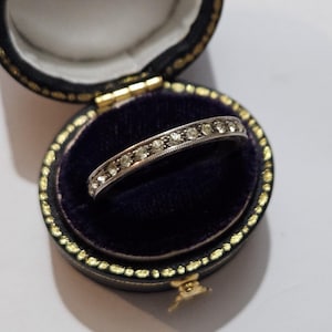 Könnte beinhalten: Ein silberner Ring mit kleinen, klaren Edelsteinen in einer Reihe, präsentiert in einer schwarzen Vintage-Schmuckschatulle mit goldenem Rand und dunkelblauem Samt. Der Ring ist der Blickfang.