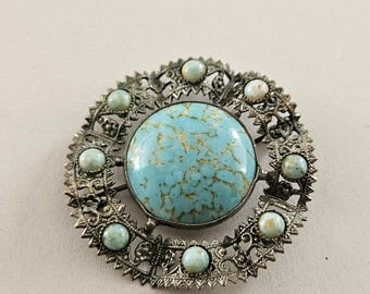 May include: Vintage silver-tone brooch with a large, round, turquoise-colored center stone. The brooch features a detailed, openwork design with smaller, light blue stones encircling the center. A classic piece of jewelry.