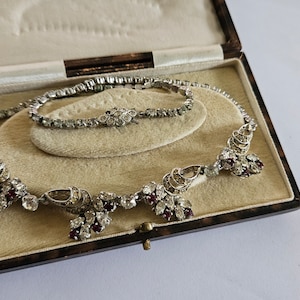 Könnte beinhalten: Ein Vintage-Rhinestone-Halsketten- und Armband-Set in einer braunen Samtbox. Die Halskette hat ein zartes Design mit roten und klaren Steinen. Das Armband ist ein schlichtes, einreihiges Design.