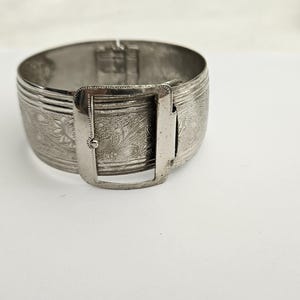 May include: A silver cuff bracelet with an engraved floral design and a rectangular clasp. The wide band is approximately 5 cm wide. The bracelet is set against a white background.
