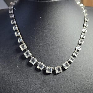Könnte beinhalten: Eine silberfarbene Halskette mit einer durchgehenden Reihe aus klaren, quadratisch geschliffenen Steinen, die in einzelnen Metallfassungen sitzen. Die Steine sind zu einer flexiblen Kette verbunden, die eine gleichmäßige, gebogene Form bildet.