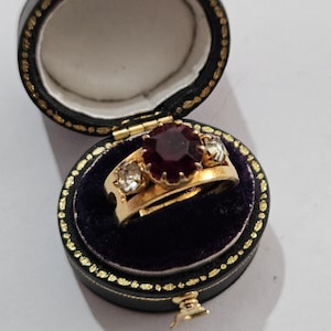 Könnte beinhalten: Ein goldener Ring mit einem großen, runden, dunkelroten Edelstein in der Mitte, flankiert von zwei kleineren, klaren Edelsteinen. Der Ring wird in einer offenen, schwarzen Schmuckschatulle mit goldenem Rand und violettem Samt präsentiert.