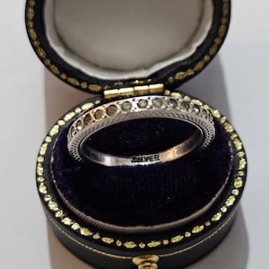 May include: A silver ring with a decorative band featuring small, round accents. The ring is inside a black jewellery box with a gold trim and a dark blue velvet interior. The word "SILVER" is stamped on the ring.