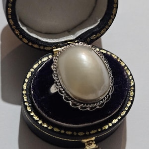 May include: A vintage ring with a large, oval, off-white pearl set in a silver filigree frame. The ring is displayed in an open, black leatherette jewelry box with a dark blue velvet interior and gold trim. The box is on a white surface.