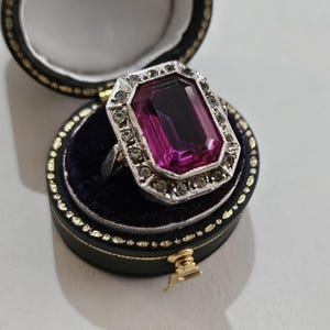 May include: A vintage ring featuring a large, rectangular, magenta gemstone set in a silver-toned metal frame, embellished with small, clear stones. The ring is presented in a black, round jewellery box with a gold clasp and trim.