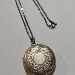 May include: A silver-toned locket necklace with a detailed floral design on the round pendant. The pendant is attached to a delicate chain. The locket is closed and has a circular, blank space in the center.