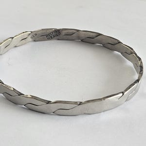 May include: A silver-coloured metal bangle bracelet with a continuous interlocking link pattern. The interior surface of the bracelet features stamped text that reads 925 and HECHO EN MEXICO, indicating the metal purity and country of origin.