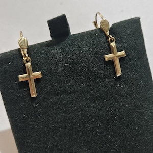 Puede incluir: Un par de pendientes de cruz dorados. Cada pendiente presenta un pequeño colgante de cruz suspendido de un gancho decorativo. Las cruces tienen un diseño simple y clásico. Los pendientes se exhiben sobre una superficie negra.