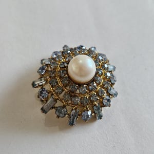 May include: A vintage-style brooch with a central faux pearl surrounded by a halo of small, light blue and clear crystals. The brooch has a gold-tone metal setting, creating a classic and elegant design.