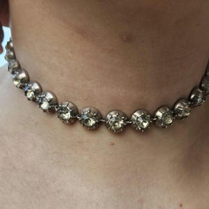 May include: A silver-toned choker necklace featuring a series of round, faceted, clear gemstones. The necklace is composed of multiple circular links, each holding a gemstone. The necklace is worn around the neck.