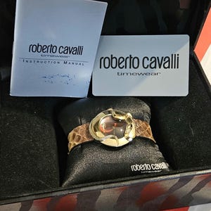 May include: A Roberto Cavalli timepiece watch with a gold-tone case and a brown snakeskin band. The watch face is a reddish-brown color. The watch is displayed in a black box with the brand name on the box and the instruction manual.