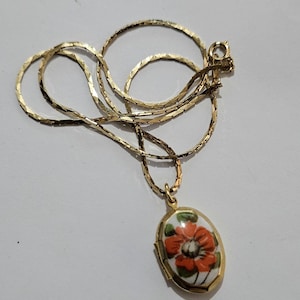 May include: A gold-toned necklace with a floral pendant. The pendant is oval-shaped, featuring a red and orange flower design on a white background. The chain has a delicate, textured pattern.