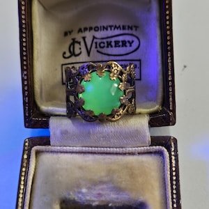 May include: A ring featuring a round, glowing green cabochon stone set in an intricate, dark metal filigree band with floral motifs, presented inside a dark brown, hinged jewelry box with the text BY APPOINTMENT J.C. VICKERY printed inside.