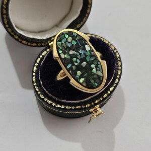 Puede incluir: Un anillo de oro con una incrustación ovalada de piezas iridiscentes verdes y negras. El anillo se muestra en un joyero negro abierto con ribete dorado y un interior de terciopelo azul oscuro. La caja está abierta, revelando el anillo.