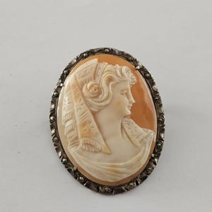 May include: An oval cameo brooch depicting a woman in profile, wearing a draped head covering. The cameo is set in a silver-coloured frame with a decorative border. The woman's face is a light peach colour, with details carved in creamy white.