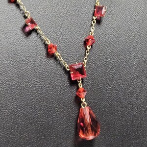May include: A gold-toned necklace featuring red gemstone accents. The necklace has a delicate chain with square and teardrop-shaped red stones. The pendant is a larger, teardrop-shaped red stone.